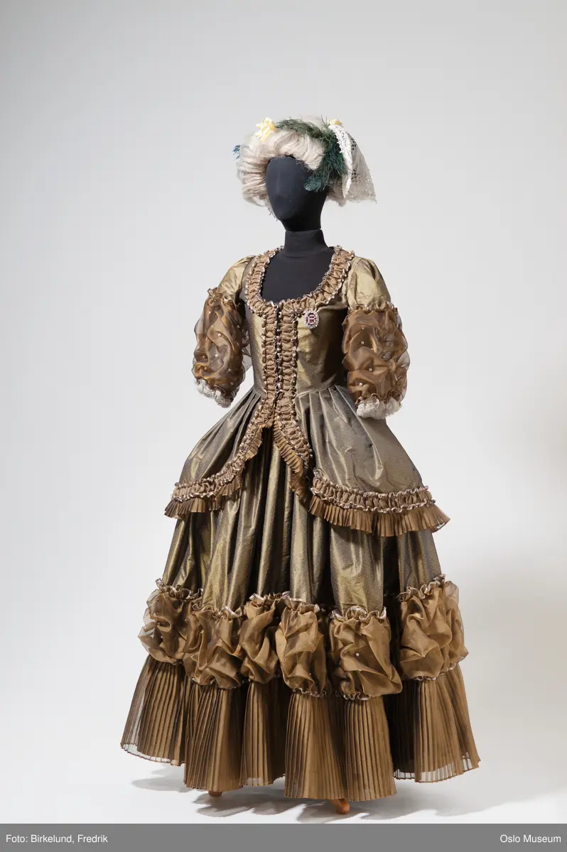 Trekvarte ermer, lang overdel, skråner ned bak, langt skjørt i grønn silke med rysjer og plisséinnslag. Lys parykk med hatt, brodert med fjær.