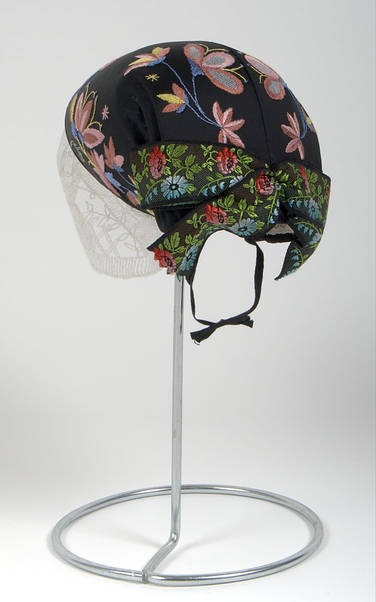 Bygdedräkt i 8 delar. 

a) Livstycke - Grönt livstycke av ylle med rosa broderier och snörning framtill.

b) Skjortlinne - Vitt skjortlinne av linne med broderier i rosa och blått. MBRD 1984.

c )Kjol - Blå handsydd kjol i ylle. Fållad med förstärkning av rödvittrutigt tyg.

d )Förkläde - Förkläde av linne. Vitt, med rosa och blåa detaljer

e) Hätta - Svart bindmössa i syntetsiden broderad med blommor i rosa, blått och gult. Nertill finns blåa och röda blommor omfamnat i ett bladverk i grönt. 

f) Sjal - Blå sjal broderad med färgrikt blommotiv.

g )Kjolväska - Grön ylleväska med rödrosa band som fåll. Broderat med M-BRD 1985. Blåvitt band för fastsättning i kjolen.

h) Kvitton - 1 st kuvert med 7st kvitton för de kostnader som dräkten gick på. Enligt givarens egna uträkningar kostade dräkten 2586kr att tillverka. Då är även kurskostnaden inräknad.

Det medföljer även fotografier på givaren iklädd dräkten och en mindre artikel om dräkten.