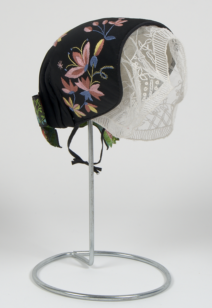 Bygdedräkt i 8 delar. 

a) Livstycke - Grönt livstycke av ylle med rosa broderier och snörning framtill.

b) Skjortlinne - Vitt skjortlinne av linne med broderier i rosa och blått. MBRD 1984.

c )Kjol - Blå handsydd kjol i ylle. Fållad med förstärkning av rödvittrutigt tyg.

d )Förkläde - Förkläde av linne. Vitt, med rosa och blåa detaljer

e) Hätta - Svart bindmössa i syntetsiden broderad med blommor i rosa, blått och gult. Nertill finns blåa och röda blommor omfamnat i ett bladverk i grönt. 

f) Sjal - Blå sjal broderad med färgrikt blommotiv.

g )Kjolväska - Grön ylleväska med rödrosa band som fåll. Broderat med M-BRD 1985. Blåvitt band för fastsättning i kjolen.

h) Kvitton - 1 st kuvert med 7st kvitton för de kostnader som dräkten gick på. Enligt givarens egna uträkningar kostade dräkten 2586kr att tillverka. Då är även kurskostnaden inräknad.

Det medföljer även fotografier på givaren iklädd dräkten och en mindre artikel om dräkten.
