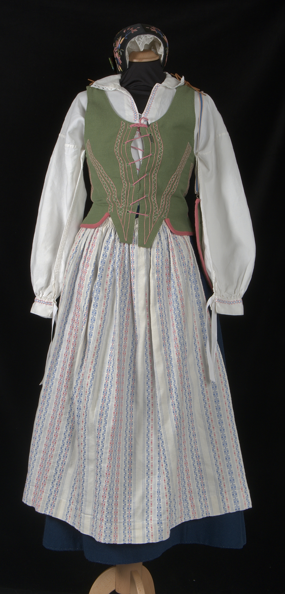 Bygdedräkt i 8 delar. 

a) Livstycke - Grönt livstycke av ylle med rosa broderier och snörning framtill.

b) Skjortlinne - Vitt skjortlinne av linne med broderier i rosa och blått. MBRD 1984.

c )Kjol - Blå handsydd kjol i ylle. Fållad med förstärkning av rödvittrutigt tyg.

d )Förkläde - Förkläde av linne. Vitt, med rosa och blåa detaljer

e) Hätta - Svart bindmössa i syntetsiden broderad med blommor i rosa, blått och gult. Nertill finns blåa och röda blommor omfamnat i ett bladverk i grönt. 

f) Sjal - Blå sjal broderad med färgrikt blommotiv.

g )Kjolväska - Grön ylleväska med rödrosa band som fåll. Broderat med M-BRD 1985. Blåvitt band för fastsättning i kjolen.

h) Kvitton - 1 st kuvert med 7st kvitton för de kostnader som dräkten gick på. Enligt givarens egna uträkningar kostade dräkten 2586kr att tillverka. Då är även kurskostnaden inräknad.

Det medföljer även fotografier på givaren iklädd dräkten och en mindre artikel om dräkten.
