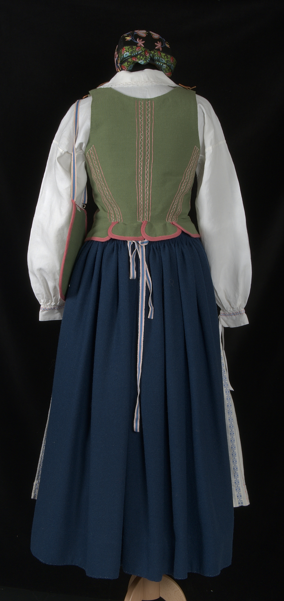 Bygdedräkt i 8 delar. 

a) Livstycke - Grönt livstycke av ylle med rosa broderier och snörning framtill.

b) Skjortlinne - Vitt skjortlinne av linne med broderier i rosa och blått. MBRD 1984.

c )Kjol - Blå handsydd kjol i ylle. Fållad med förstärkning av rödvittrutigt tyg.

d )Förkläde - Förkläde av linne. Vitt, med rosa och blåa detaljer

e) Hätta - Svart bindmössa i syntetsiden broderad med blommor i rosa, blått och gult. Nertill finns blåa och röda blommor omfamnat i ett bladverk i grönt. 

f) Sjal - Blå sjal broderad med färgrikt blommotiv.

g )Kjolväska - Grön ylleväska med rödrosa band som fåll. Broderat med M-BRD 1985. Blåvitt band för fastsättning i kjolen.

h) Kvitton - 1 st kuvert med 7st kvitton för de kostnader som dräkten gick på. Enligt givarens egna uträkningar kostade dräkten 2586kr att tillverka. Då är även kurskostnaden inräknad.

Det medföljer även fotografier på givaren iklädd dräkten och en mindre artikel om dräkten.