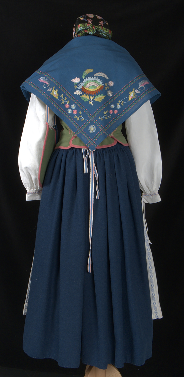 Bygdedräkt i 8 delar. 

a) Livstycke - Grönt livstycke av ylle med rosa broderier och snörning framtill.

b) Skjortlinne - Vitt skjortlinne av linne med broderier i rosa och blått. MBRD 1984.

c )Kjol - Blå handsydd kjol i ylle. Fållad med förstärkning av rödvittrutigt tyg.

d )Förkläde - Förkläde av linne. Vitt, med rosa och blåa detaljer

e) Hätta - Svart bindmössa i syntetsiden broderad med blommor i rosa, blått och gult. Nertill finns blåa och röda blommor omfamnat i ett bladverk i grönt. 

f) Sjal - Blå sjal broderad med färgrikt blommotiv.

g )Kjolväska - Grön ylleväska med rödrosa band som fåll. Broderat med M-BRD 1985. Blåvitt band för fastsättning i kjolen.

h) Kvitton - 1 st kuvert med 7st kvitton för de kostnader som dräkten gick på. Enligt givarens egna uträkningar kostade dräkten 2586kr att tillverka. Då är även kurskostnaden inräknad.

Det medföljer även fotografier på givaren iklädd dräkten och en mindre artikel om dräkten.
