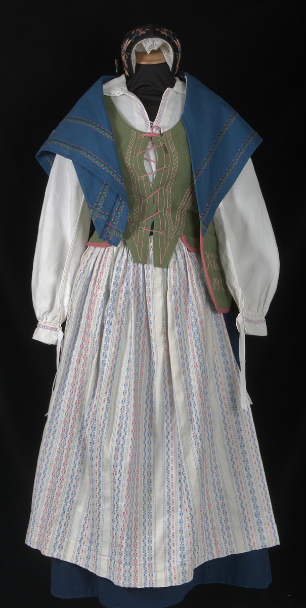 Bygdedräkt i 8 delar. 

a) Livstycke - Grönt livstycke av ylle med rosa broderier och snörning framtill.

b) Skjortlinne - Vitt skjortlinne av linne med broderier i rosa och blått. MBRD 1984.

c )Kjol - Blå handsydd kjol i ylle. Fållad med förstärkning av rödvittrutigt tyg.

d )Förkläde - Förkläde av linne. Vitt, med rosa och blåa detaljer

e) Hätta - Svart bindmössa i syntetsiden broderad med blommor i rosa, blått och gult. Nertill finns blåa och röda blommor omfamnat i ett bladverk i grönt. 

f) Sjal - Blå sjal broderad med färgrikt blommotiv.

g )Kjolväska - Grön ylleväska med rödrosa band som fåll. Broderat med M-BRD 1985. Blåvitt band för fastsättning i kjolen.

h) Kvitton - 1 st kuvert med 7st kvitton för de kostnader som dräkten gick på. Enligt givarens egna uträkningar kostade dräkten 2586kr att tillverka. Då är även kurskostnaden inräknad.

Det medföljer även fotografier på givaren iklädd dräkten och en mindre artikel om dräkten.