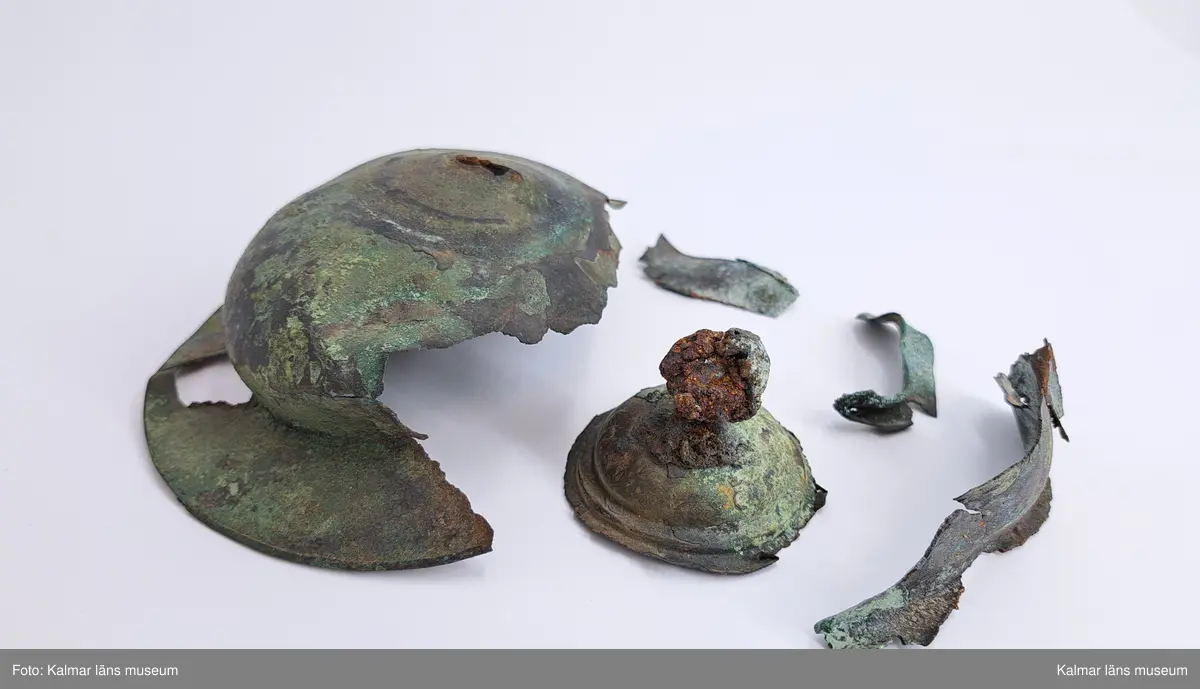 KLM 4911. Sköldbuckla, av brons. Den översta knoppen är av järn och överdragen av brons. En halvsfärisk nit. Sköldbucklan är i flera delar. Gravfynd tillsammans med KLM 4912-4915.