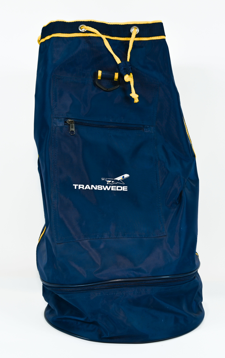 Mörkblå sjösäck med gula detaljer. På väskan står 'TRANSWEDE' och ett avbildat flygplan. Öppningen sitter upptill och går att stänga med ett gult rep samt en hake. Väskan går även att öppna från sidan genom en dragkedja. Det finns två fickor, en framtill samt en vid botten, båda med dragkedja.