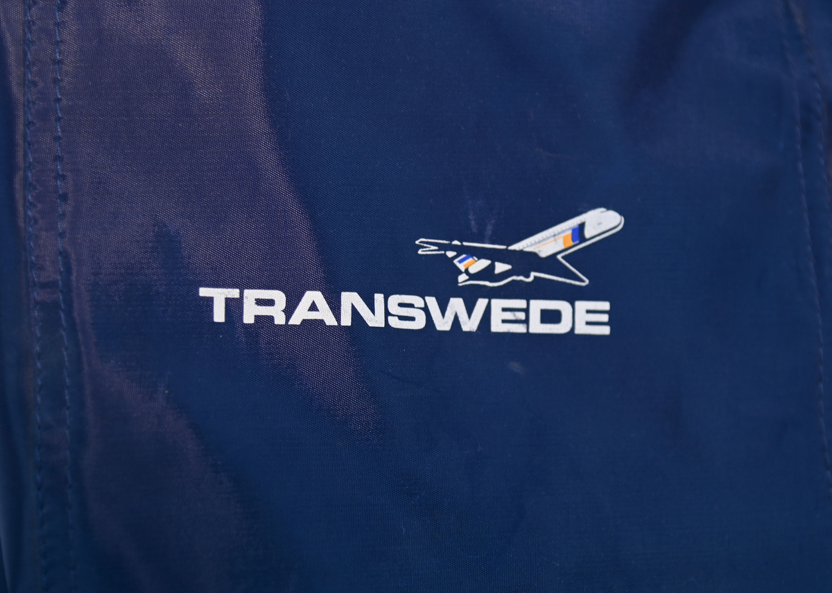 Mörkblå sjösäck med gula detaljer. På väskan står 'TRANSWEDE' och ett avbildat flygplan. Öppningen sitter upptill och går att stänga med ett gult rep samt en hake. Väskan går även att öppna från sidan genom en dragkedja. Det finns två fickor, en framtill samt en vid botten, båda med dragkedja.