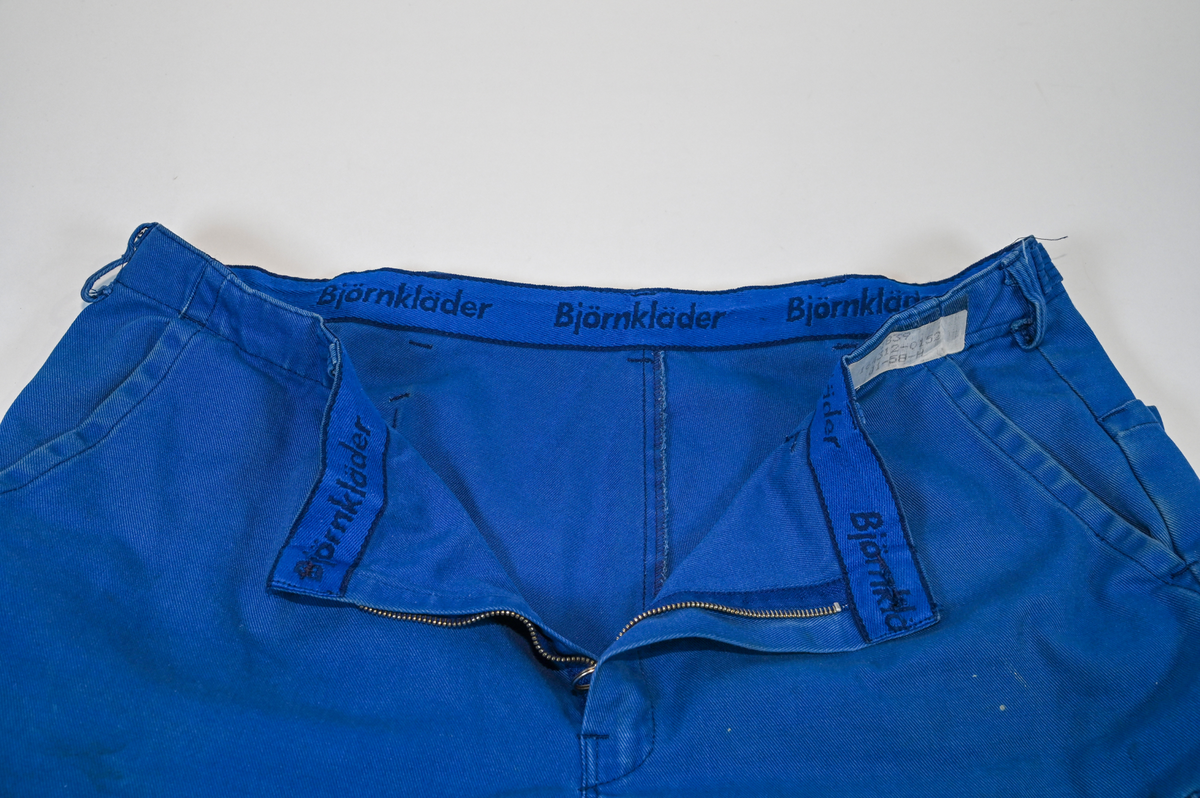Byxa i blått tyg i modell arbetsbyxa. På båda byxbenen finns invändiga fickor för knäskydd. Sju öglor för livrem samt nio fickor varav en har lock med metallknapp. På vardera sida om byxlinningen sitter en kapp med tillhörande tygbit för att kunna reglera midjan. Storlek 58.

"Björnkläder" står skrivet på en fastsydd tygetikett på vänster bakficka. Texten är skriven i vita bokstäver och bakgrunden är blå.

"Björnkläder" står även skrivet på insidan längs med byxlinningen. Texten är skriven i mörkblåa bokstäver på blå bakgrund.

"Björnkläder / 58" står på fastsydd etikett vid inre byxlinning tillsammans med motiv av en björn. Texten är i gult på blå bakgrund.