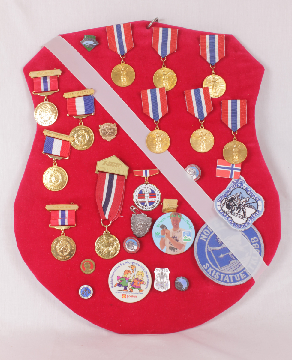Skjoldformet plate med idrettsmerker. Merkene er medaljer, buttons, pins og strykemerker.