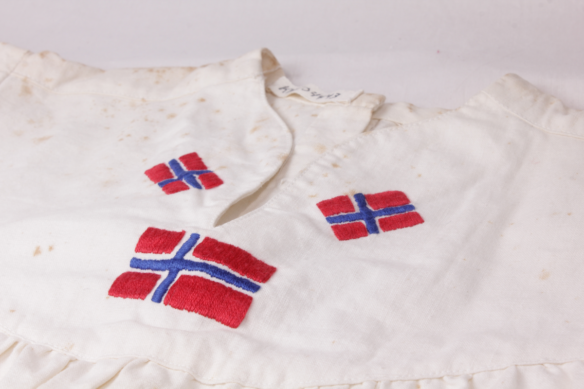 Hvit 17. mai-kjole fra 1945. Sydd av blekede sukkersekker eller melsekker og brodert med norske flagg. Kjolen har 12 falske knapper på rygg og lukkes med seks skjulte trykknapper. Glidelås i siden.