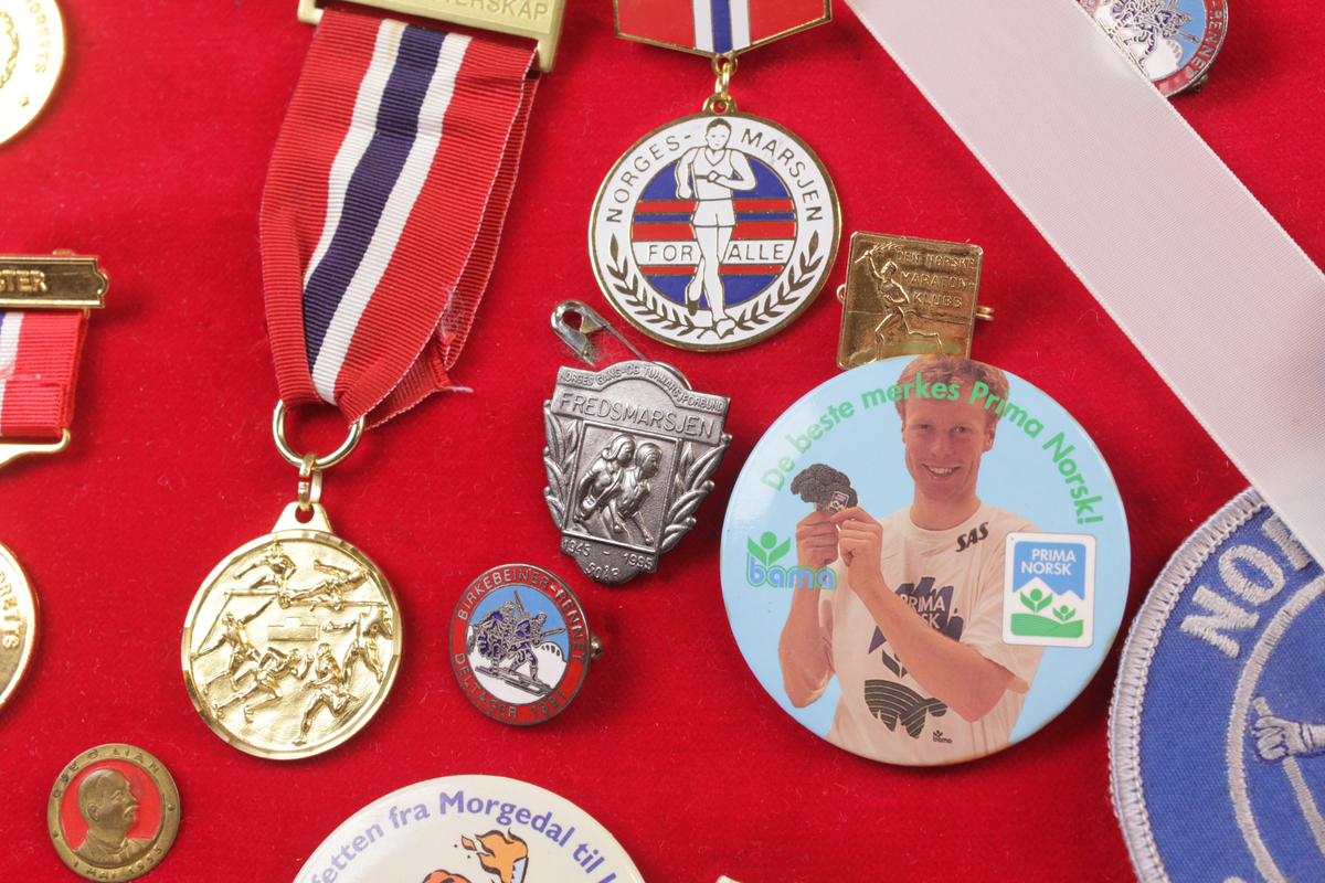 Skjoldformet plate med idrettsmerker. Merkene er medaljer, buttons, pins og strykemerker.