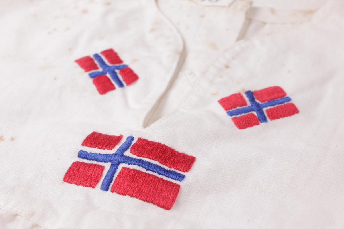 Hvit 17. mai-kjole fra 1945. Sydd av blekede sukkersekker eller melsekker og brodert med norske flagg. Kjolen har 12 falske knapper på rygg og lukkes med seks skjulte trykknapper. Glidelås i siden.