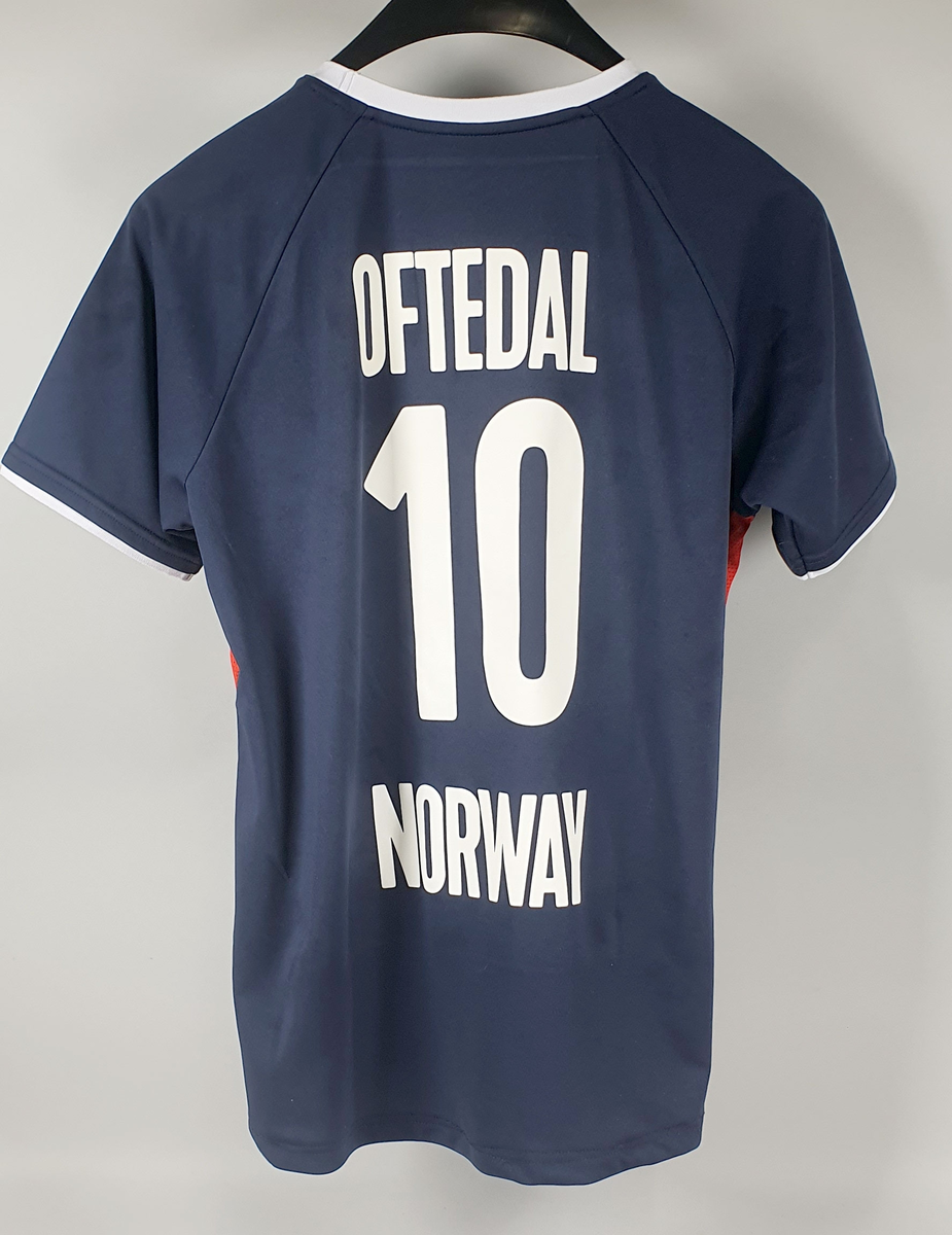 Mørkeblå håndballdrakt bestående av t-skjorte, shorts og svettebånd til håndledd. T-skjorten har hvite kantbånd i halsen og ytterst på ermene, en rød stripe i sidene, og et norsk flagg i en hvit ring på brystet. Buksa har knytebånd i livet og norsk flagg i en hvit ring nederst på venstre side, og tallet 10 nederst på høyre side foran.