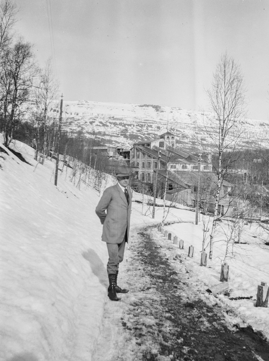 Pent kledd ukjent mann i vinterlandskap. Oppredningsverket ved Bjørkåsen gruver i bakgrunnen.