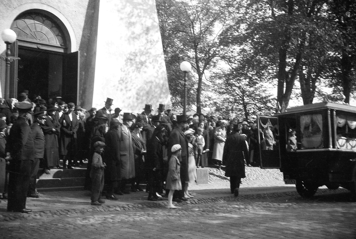 Begravningsprocession med bilar och hästar utanför Varbergs kyrka. Gatuvy från Kungsgatan. Bild 1: Hästekipage med kistan täckt av vita liljor. Kusken kan vara Alvar på Träslövsvägen som med sina hästar brukade köra vid begravningar. I förgrunden ses en prestaver. Sett från söder. Bild 2: De sörjande kommer ut från kyrkan och går mot likbilen till höger. Bild 3: Processionen sedd från norr med två hästekipage och en öppen bil.