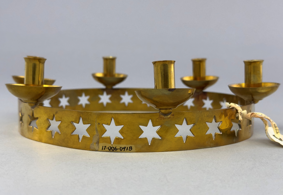 Ljusstake, "Luciakrona" för sex små julgransljus, gjord av mässingband med utstansat stjärnmönster. Ljushållarna är fastlödda på bandets högkant.
