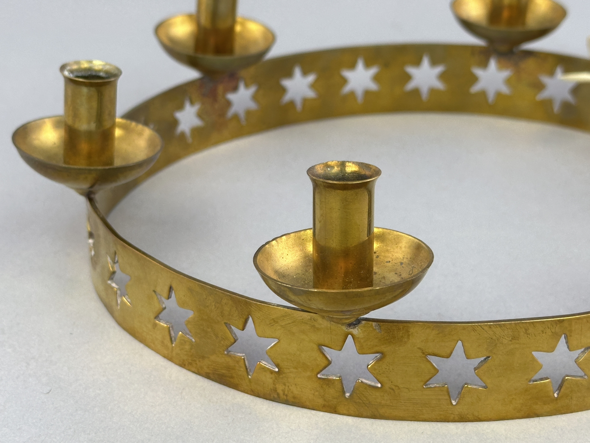 Ljusstake, "Luciakrona" för sex små julgransljus, gjord av mässingband med utstansat stjärnmönster. Ljushållarna är fastlödda på bandets högkant.