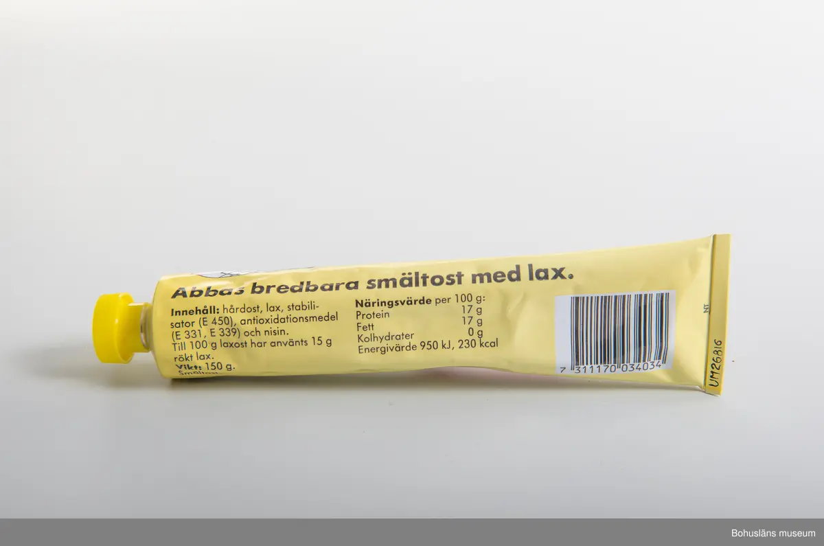 Tryckt dekor i gult,rosa,svart,vitt Gul gjuten plasthatt. Text: "Abbas Lax-Ost. Nyhet  samt produktbild  På baksidan innehållsförteckning och prismärkningskod. 
Tuben är identisk med UM026808
Om givaren se UM026667