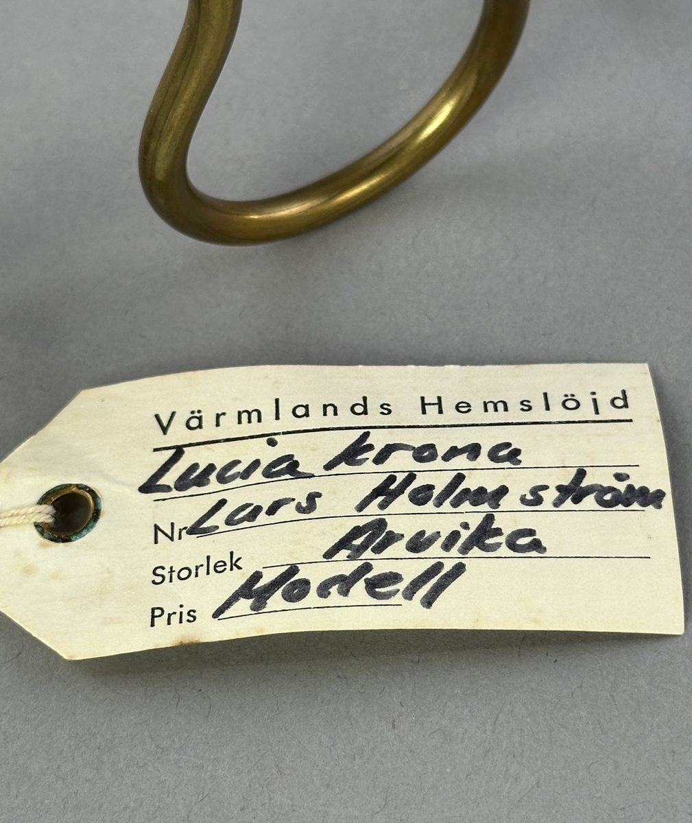 Ljusstake, "Luciakrona" för sju små stearinljus, gjord av mässingtråd som böjts i mjuk vågform. Högst upp på varje böj sitter en ljushållare fastnitad.