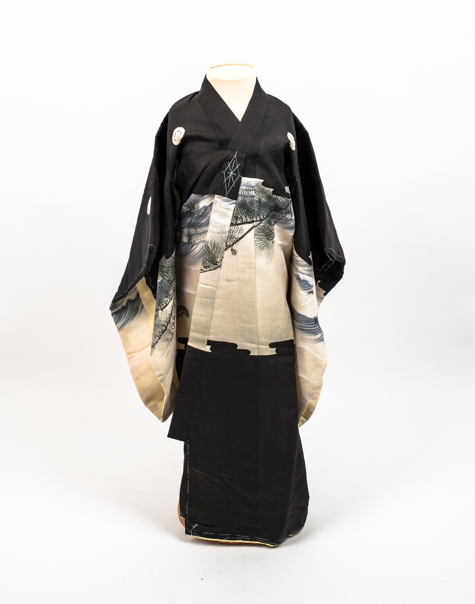 Barnekimono i svart silke med kremfarget parti med figur- og landskapsmotiv med blant annet traner, to gamle menn og broderte kongler. Kimonoen er delvis vattert og fôret med gul silke. Antagelig til jente.