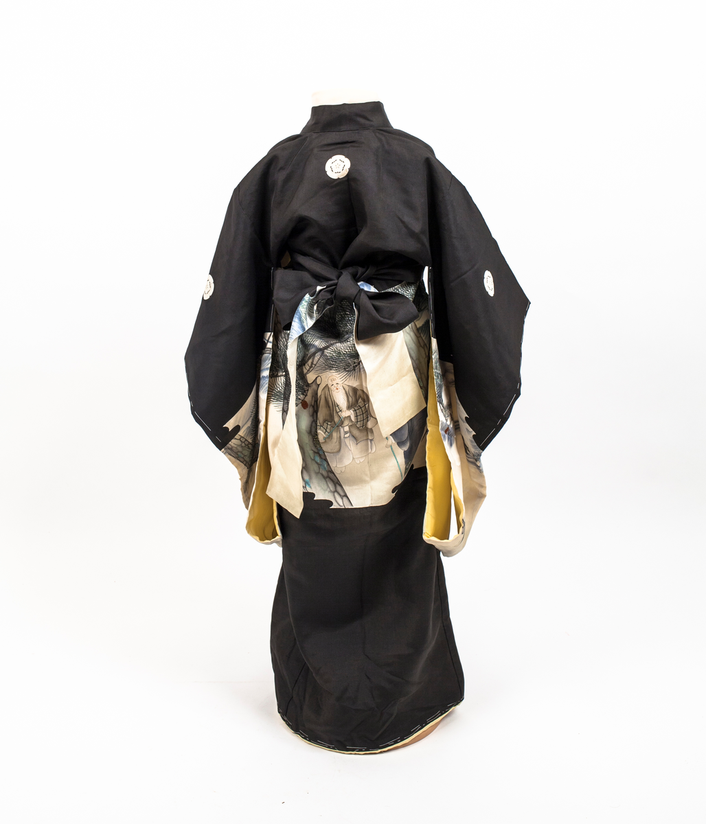 Barnekimono i svart silke med kremfarget parti med figur- og landskapsmotiv med blant annet traner, to gamle menn og broderte kongler. Kimonoen er delvis vattert og fôret med gul silke. Antagelig til jente.