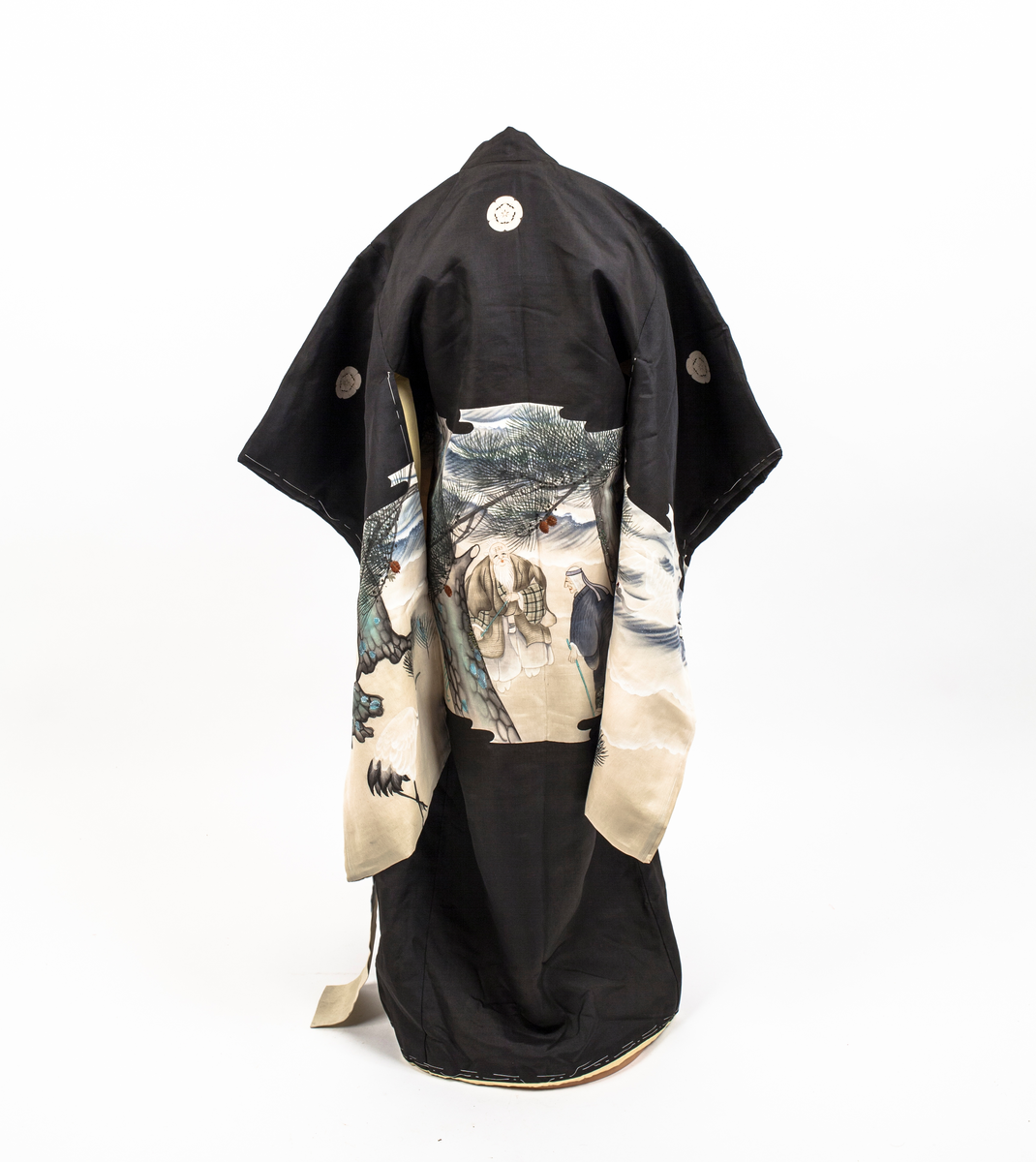 Barnekimono i svart silke med kremfarget parti med figur- og landskapsmotiv med blant annet traner, to gamle menn og broderte kongler. Kimonoen er delvis vattert og fôret med gul silke. Antagelig til jente.
