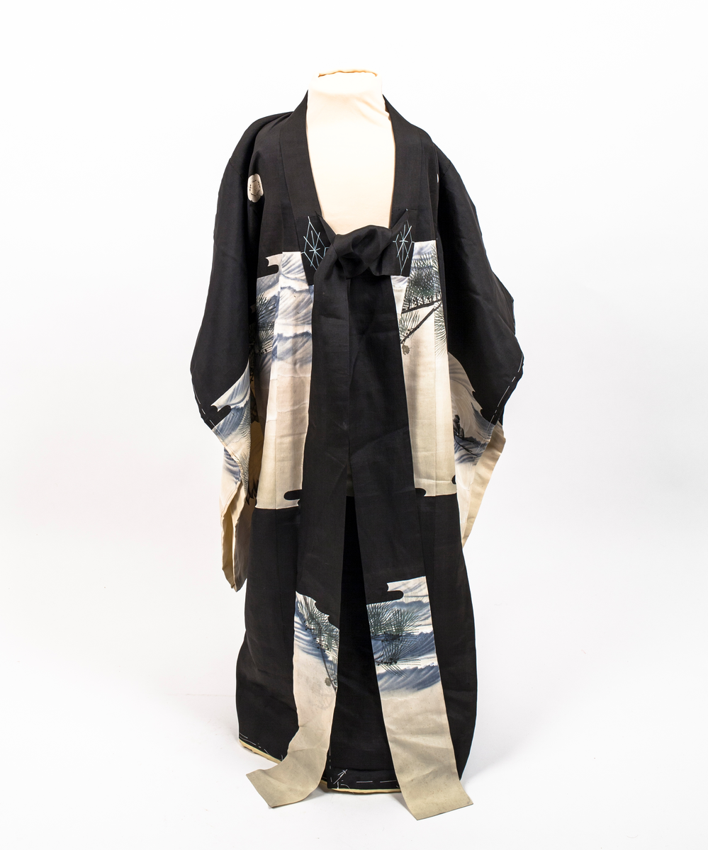 Barnekimono i svart silke med kremfarget parti med figur- og landskapsmotiv med blant annet traner, to gamle menn og broderte kongler. Kimonoen er delvis vattert og fôret med gul silke. Antagelig til jente.