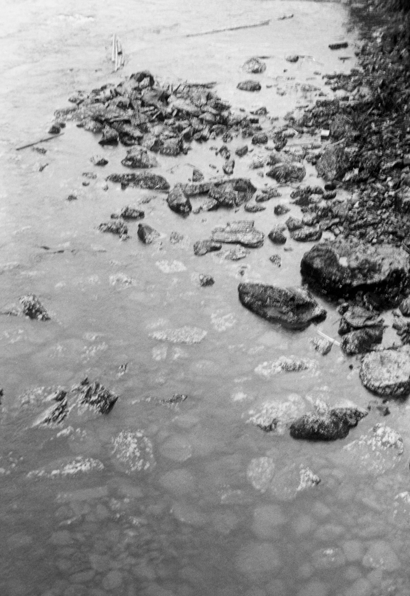 Detaljbilde av elvetopografien i Gudbrandsdalslågen, like nedenfor Hunderfossen, sommeren 1961. Fotografiet er tatt fra en av pilarene på motstrøms side av den østre enden av jernbanebrua, med kameraet vendt nedover mot vannet og den elvebotnen. Her var det grunt og steinete, og det innebar en åpenbar fare for at fløtingsvirket kunne sette seg fast og binde seg i digre hauger eller vaser.

Dette fotografiet ble tatt i 1961, på et tidspunkt da bygginga av elvekraftverket ved den ovenforliggende Hunderfossen var i gang, antakelig for å dokumentere fløtingsforholdene før reguleringsprosjektet ble realisert. En tilsvarende bildeserie ble også tatt i 1959, men på et tidspunkt med litt annen vannføring. Forholdene da dette fotografiet ble tatt beskrives slik i en tekst i et av Glomma fellesfløtingsforenings album: «Notater fra Befaring, Lågen, Isakstuøren-Hunderfos 3/8-61 med vannføring 403 m3/sek., målt i Losna. (Ment som supplement til Hollums forutgående undersøkelser på vannføring 252 m3/sek. – foregående sider).» I Glomma fellesfløtingsforenings album er dette motivet tekstet slik: «Ør nedenfor j. b. broen har vi også i form av et storstenet grunt område ovenfor broen. Tatt over pillar på solsiden».