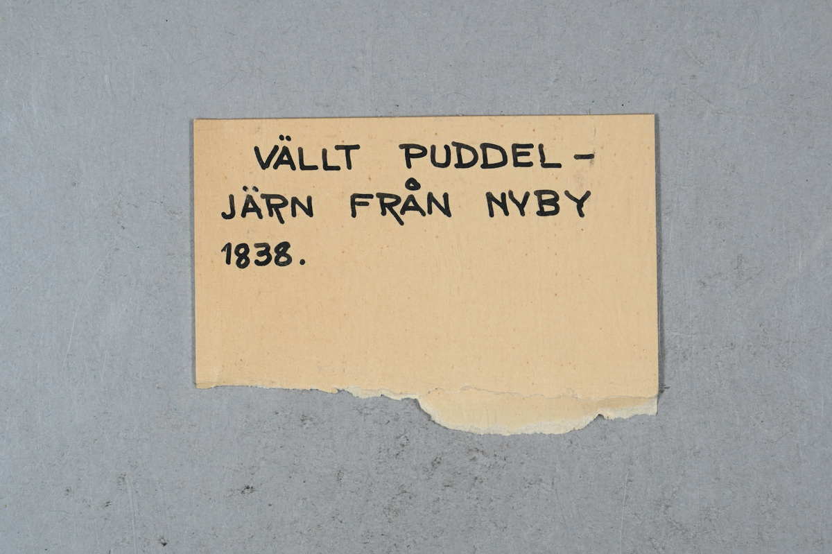 Prov på vällt puddeljärn. Prov taget från Nyby år 1838. Etikett på föremålet: "Wälld Puddlingssmälta af 73% Puddlingsugnen, Rodgers skift, Tackjernet OOL (?). 25. Nyby den 27 Apr. 1838." Tillhörande lapp med notering: "Vällt puddeljärn från Nyby 1838."
