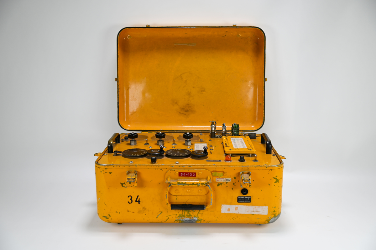 Gul plåtlåda med lock på gångjärn. Under locket en panel med 5 rattar och 3 inglasade mätare och 2 vevar. Anslutnings uttag där slangar ska skruvas fast i maskinen. Påskrift på utsidan: ”Garrett Manufacturin limited” ”Made in Canada”, i locket finns en fasttejpad kopia av en text med rubriken: ”Warning! This machine is subject to brakedownes during periods of crititcal need”, texten är ett skämt och uppmanar observatören att ”keep cool and say nice things to the machine”.