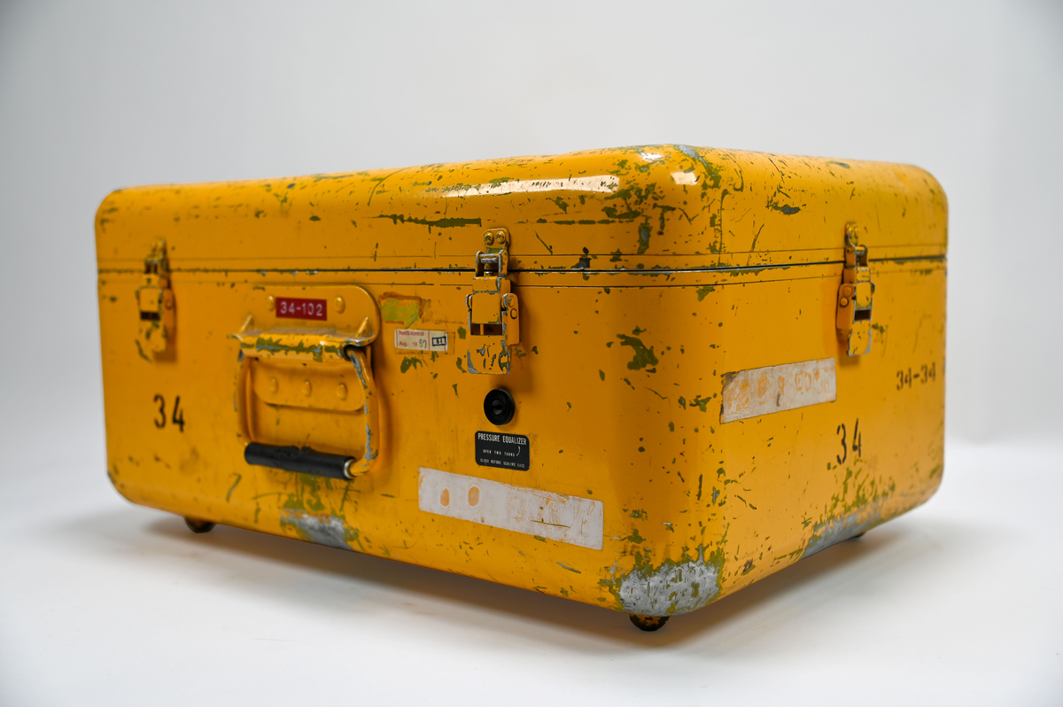 Gul plåtlåda med lock på gångjärn. Under locket en panel med 5 rattar och 3 inglasade mätare och 2 vevar. Anslutnings uttag där slangar ska skruvas fast i maskinen. Påskrift på utsidan: ”Garrett Manufacturin limited” ”Made in Canada”, i locket finns en fasttejpad kopia av en text med rubriken: ”Warning! This machine is subject to brakedownes during periods of crititcal need”, texten är ett skämt och uppmanar observatören att ”keep cool and say nice things to the machine”.