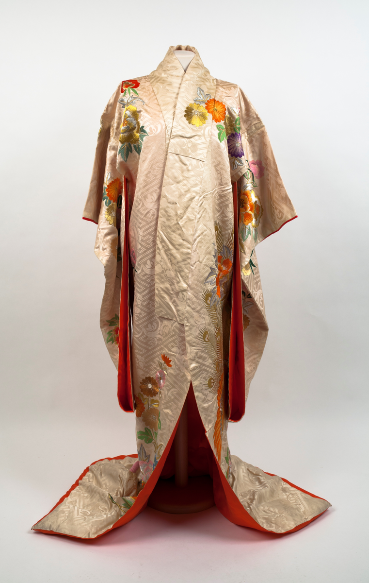 Brudekimono i lys beige silkesateng rikt dekorert med mønster av fugler, blomster, bladverk og geometriske streker. På venstre side en påfugl brodert i orange og gull sammen med store blomster i mange farger og bladverk. Påfuglen blir tatt igjen på høyre skulder bak. Dekor på ermene og foran på hvert forstykke. Vattert i nedre kant og sydd slik at fôret som er oransje er synlig på retta.
