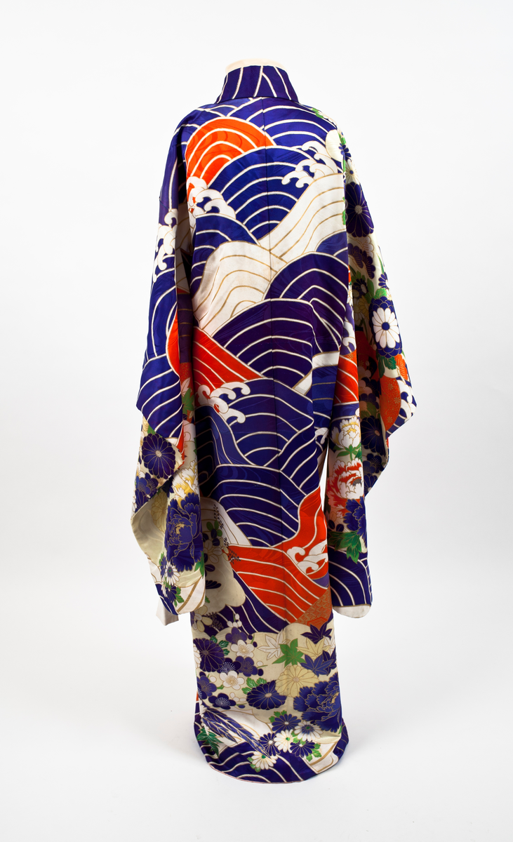 Kimono i mønstret silke. Stort bølgemønster i blått, hvitt og rødt med gullfargede kanter. I kimonoens nedre del og på ermene blomstermønster med krysantemer og peoner mot gullfarget bunn. Påsydde hvite bånd som kan knytes.
