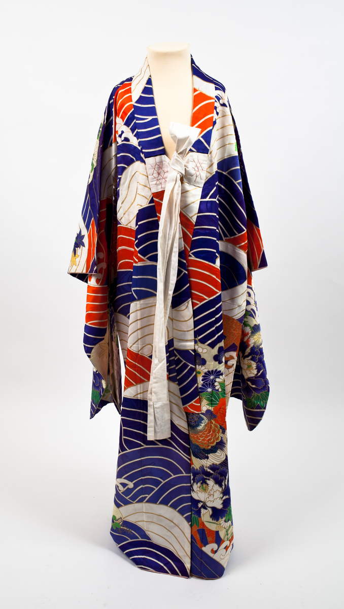 Kimono i mønstret silke. Stort bølgemønster i blått, hvitt og rødt med gullfargede kanter. I kimonoens nedre del og på ermene blomstermønster med krysantemer og peoner mot gullfarget bunn. Påsydde hvite bånd som kan knytes.