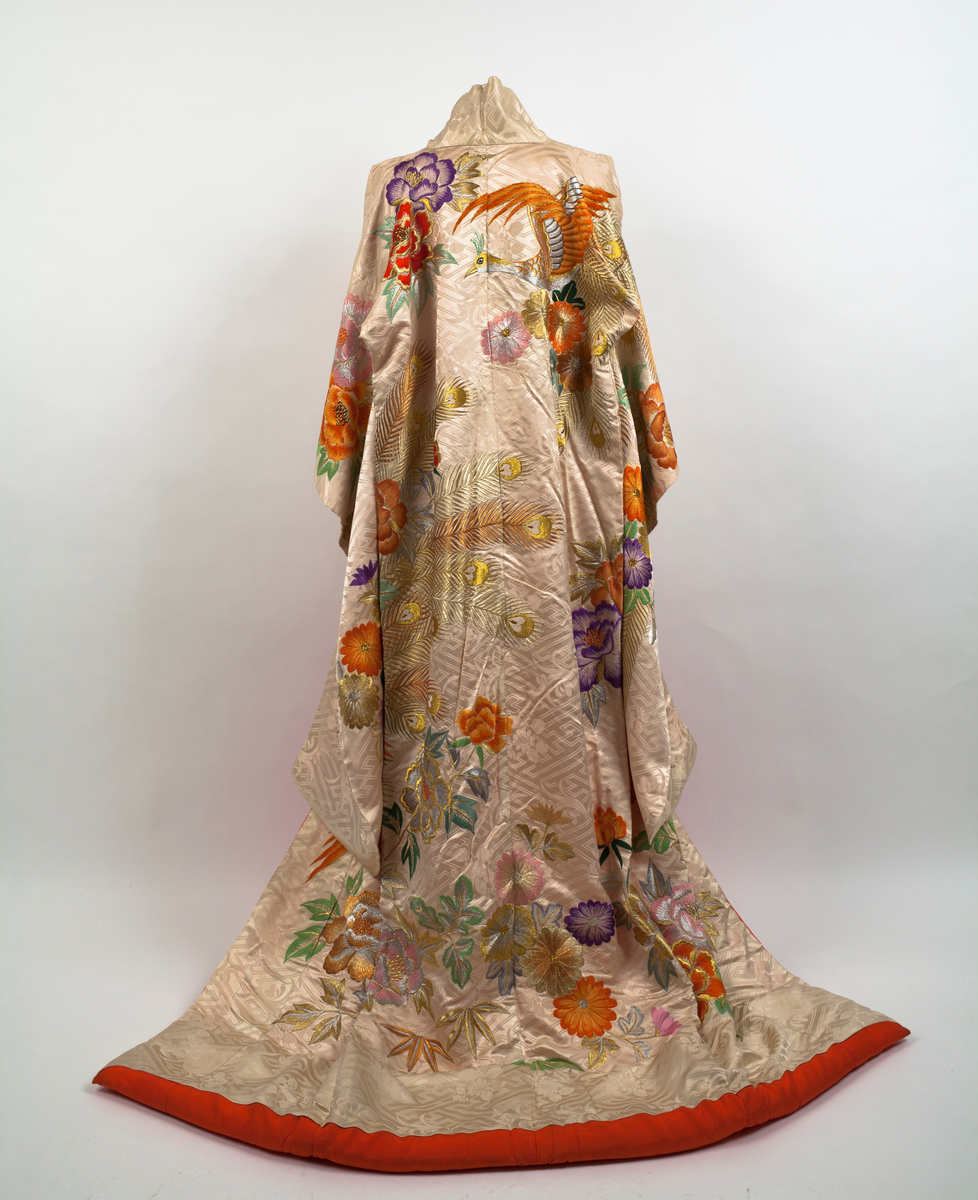 Brudekimono i lys beige silkesateng rikt dekorert med mønster av fugler, blomster, bladverk og geometriske streker. På venstre side en påfugl brodert i orange og gull sammen med store blomster i mange farger og bladverk. Påfuglen blir tatt igjen på høyre skulder bak. Dekor på ermene og foran på hvert forstykke. Vattert i nedre kant og sydd slik at fôret som er oransje er synlig på retta.