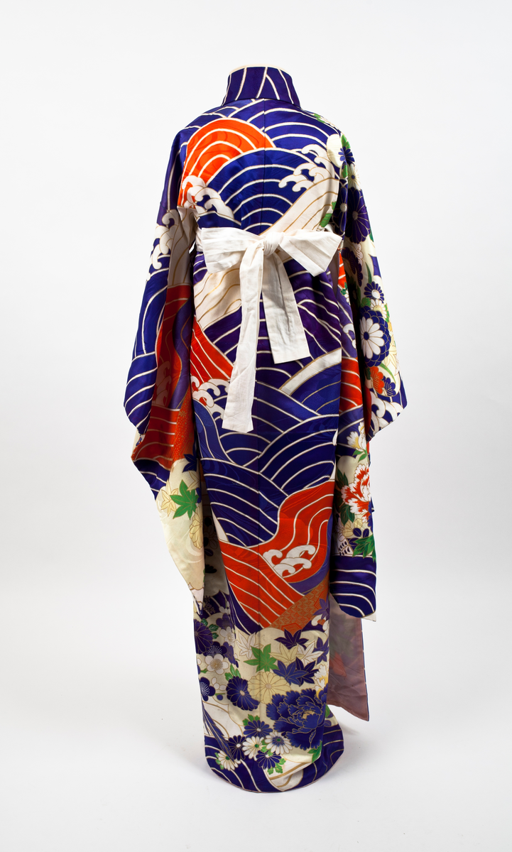 Kimono i mønstret silke. Stort bølgemønster i blått, hvitt og rødt med gullfargede kanter. I kimonoens nedre del og på ermene blomstermønster med krysantemer og peoner mot gullfarget bunn. Påsydde hvite bånd som kan knytes.