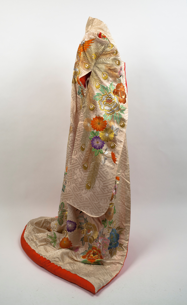 Brudekimono i lys beige silkesateng rikt dekorert med mønster av fugler, blomster, bladverk og geometriske streker. På venstre side en påfugl brodert i orange og gull sammen med store blomster i mange farger og bladverk. Påfuglen blir tatt igjen på høyre skulder bak. Dekor på ermene og foran på hvert forstykke. Vattert i nedre kant og sydd slik at fôret som er oransje er synlig på retta.