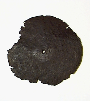 En i det närmaste rund (kanterna ojämna, anfrätta av korrosion) järnplatta. Något konisk (svag utbuktning i mitten). Ca 1 mm tjock i kanterna, ca 2 mm i centrum. I mitten ett ca 7 mm stort hål. Enl. tidigare anteckningar (se bild nr 2) har stycket tidigare varit försett med ett skaft, vilket numera är förkommet.
