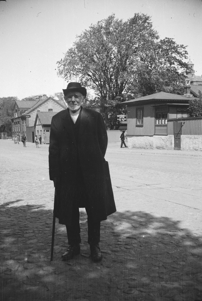Gatuvy från Västra Vallgatan i Varberg, två bilder. Porträtt av en man, möjligen "Mauritz i Sp" (notering i negativalbum), där Sp står för Spannarp. Han var en slaktare som stod på torget med sina varor. Mannen är klädd i överrock och hatt. På den lilla byggnaden till höger på bild 1 sitter en butiksskylt för "SKRÄDDERI AFFÄR KLAS GLANS", kv Gästgivaren. Bebyggelsen bakom butiken ligger i kv Läkaren. Mellan kvarteren går Bäckgatan och på bild 2 ser man hörnen av Gerlachska huset samt prästgården närmast i bild.