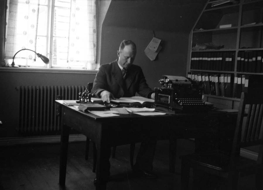 Manlig kontorist sitter vid ett skrivbord med en bok. Bredvid honom står ett stämpelställ och en skrivmaskin är vänd mot bordets andra sida. På skrivbordet står en skrivmaskin och i hyllan längs väggen förvaras pärmar. Bild 2 visar mannen i närbild.