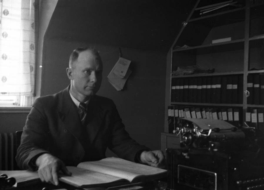 Manlig kontorist sitter vid ett skrivbord med en bok. Bredvid honom står ett stämpelställ och en skrivmaskin är vänd mot bordets andra sida. På skrivbordet står en skrivmaskin och i hyllan längs väggen förvaras pärmar. Bild 2 visar mannen i närbild.