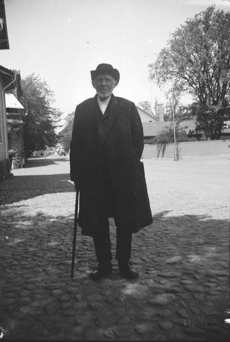 Gatuvy från Västra Vallgatan i Varberg, två bilder. Porträtt av en man, möjligen "Mauritz i Sp" (notering i negativalbum), där Sp står för Spannarp. Han var en slaktare som stod på torget med sina varor. Mannen är klädd i överrock och hatt. På den lilla byggnaden till höger på bild 1 sitter en butiksskylt för "SKRÄDDERI AFFÄR KLAS GLANS", kv Gästgivaren. Bebyggelsen bakom butiken ligger i kv Läkaren. Mellan kvarteren går Bäckgatan och på bild 2 ser man hörnen av Gerlachska huset samt prästgården närmast i bild.