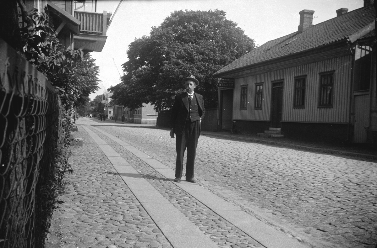 Gatuvy från Drottninggatan i Varberg där en man står vid ett trähus i kv Krögaren till höger. På huset sitter en reklamskylt för en målare ovanför portlidret. I bakgrunden ses Rådhuset vid torget, som ligger till vänster ur bild. Huset till vänster ligger i kv Sparbanken.
