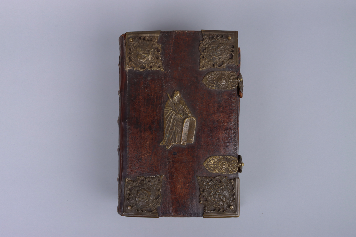 Bibel, marmorerat skinnband med upphöjda bind, dekorerad rygg. På framsida beslag i genombruten mässing med olika apostlar mitt på Moses med stentavlor. Omtag och lås med Karl XII monogram. Bibeln rymmer flertalet planscher och porträtt samt utvik med kartor.