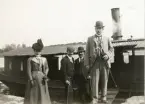 Ett sällskap med tre män och en kvinna med hatt framför en ångslup, Gysinge omkring 1900.