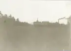 Öppet skogsfält i förgrunden och bruksvy i bakgrunden med skorstensrök från sulfitfabriken i Gysinge.
