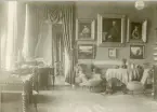 Foto visar ett rum med bl.a soffa, fåtöljer, bord, stora målningar med breda ramar, draperi mm. inifrån Gysinge herrgård.