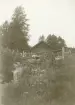 Liten stuga/uthus med gärdsgård och vedtrave omgiven av skog, troligen i trakterna runt Gysinge.