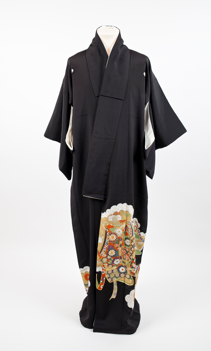 Kimono i svart silkekrepp med påtrykt orientalsk mønster plassert diagonalt på nedre del av plagget; skylignende felt med blomster og blader i hvitt, fiolett, gult, rustrød og grønnyanser, konturert med sølv- og gull-lisser. Kimonoen har dobbel krage og er fôret med hvit silkekrepp. Små ornamenter (komon) plassert i nakken, på skuldre og ermer.