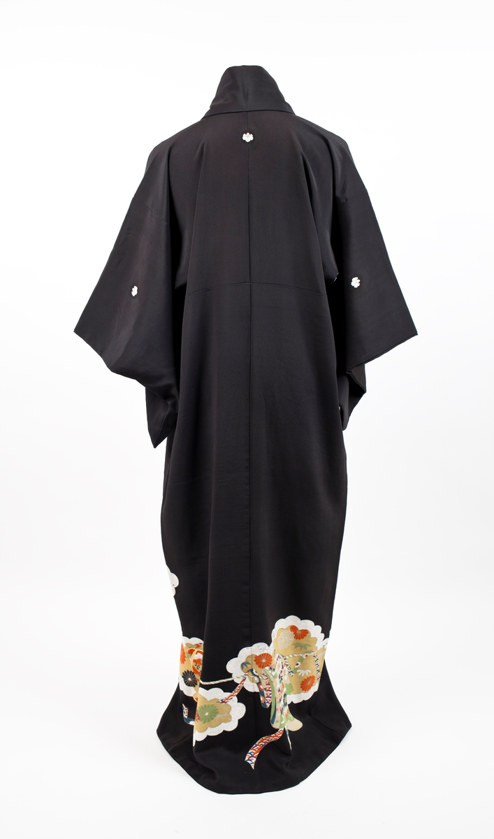 Kimono i svart silkekrepp med påtrykt orientalsk mønster plassert diagonalt på nedre del av plagget; skylignende felt med blomster og blader i hvitt, fiolett, gult, rustrød og grønnyanser, konturert med sølv- og gull-lisser. Kimonoen har dobbel krage og er fôret med hvit silkekrepp. Små ornamenter (komon) plassert i nakken, på skuldre og ermer.