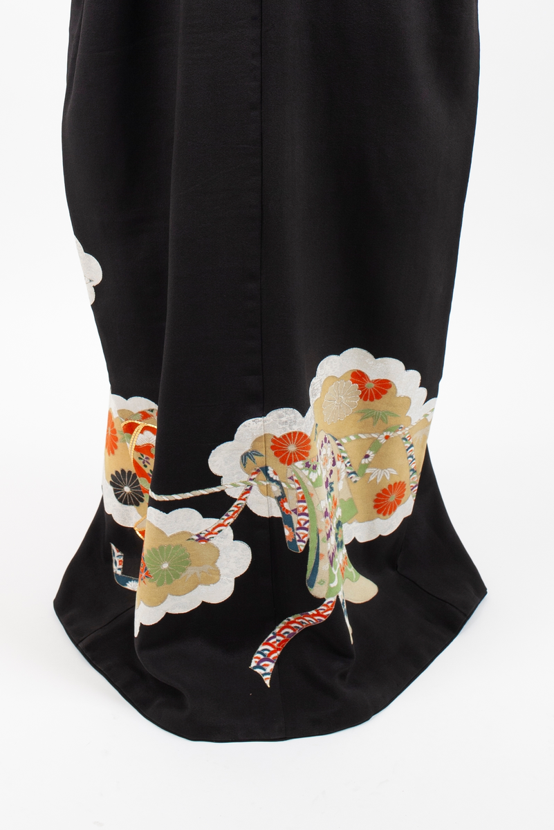 Kimono i svart silkekrepp med påtrykt orientalsk mønster plassert diagonalt på nedre del av plagget; skylignende felt med blomster og blader i hvitt, fiolett, gult, rustrød og grønnyanser, konturert med sølv- og gull-lisser. Kimonoen har dobbel krage og er fôret med hvit silkekrepp. Små ornamenter (komon) plassert i nakken, på skuldre og ermer.